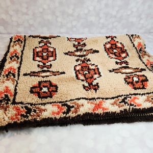May include: A beige rug with a repeating pattern of brown and orange floral designs. The rug has a dark brown fringe and border with orange and white accents. The rug is made of a soft, textured material.