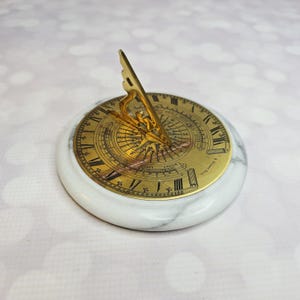 May include: A vintage sundial with a gold-colored face and a marble base. The sundial features Roman numerals and intricate markings. The gnomon is a gold-colored triangle. A decorative item for home or office.