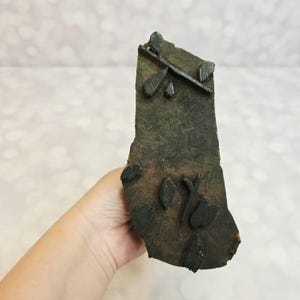 May include: A dark brown, weathered wooden block print featuring a raised, carved design of small leaves and thin stems. The surface of the block shows natural wood grain and texture, with the carved elements standing out in relief for printing.