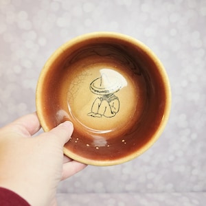 May include: A small, round ceramic bowl with a brown exterior and a tan interior. The bowl features a hand-drawn illustration of a person wearing a sombrero. The bowl is held by a hand against a blurred background.