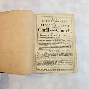 Puede incluir: Un libro antiguo abierto con el título "The Intercourses of Divine Love" impreso en la página derecha. El texto está en tinta negra sobre papel envejecido de color blanquecino. Las páginas del libro están ligeramente amarillentas.