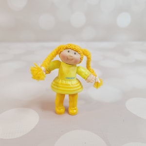 May include: A vintage Cabbage Patch Kids doll figurine. The doll has yellow hair in pigtails and is wearing a yellow dress with a striped skirt. The doll's arms are outstretched, and it has a smiling face.