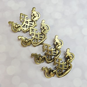 May include: Four antique brass drawer pulls with an ornate, symmetrical design. Each pull features a curved handle and a decorative openwork pattern. The brass hardware has a warm, aged finish, suggesting vintage or retro style. Perfect for furniture restoration projects.