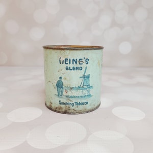 May include: A vintage, light blue metal tin with a white label. The label features the text "Meine's Blend" and a black and white illustration of a windmill and a person in traditional Dutch clothing. The text "Smoking Tobacco" is printed below the illustration.
