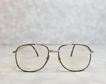 Puede incluir: Gafas de estilo aviador con una montura fina de metal en tono dorado y un diseño de doble puente. Las lentes son transparentes y tienen una línea horizontal visible que indica una función bifocal. Las varillas tienen un patrón de carey.