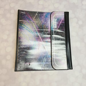May include: A Mead Trapper Keeper binder featuring a dark, cosmic-inspired design with a geometric triangular grid overlay, shimmering light streaks in blue, purple, and white, and a black vertical spine with a fold-over magnetic closure flap.