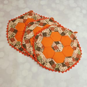 May include: Three quilted pot holders featuring a hexagonal pattern of solid orange fabric and floral print fabric in shades of brown, green, and cream. Each pot holder has a scalloped orange fabric edge and a small fabric hanging loop.
