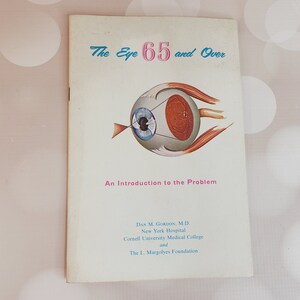 Puede incluir: Portada de libro vintage titulada "The Eye 65 and Over" con una ilustración de un ojo. La portada es blanquecina con texto azul y rosa. El libro es una introducción al problema de la salud ocular.