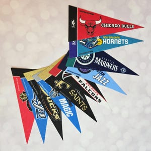 May include: A selection of pennant flags displaying logos of various sports teams. The triangular flags showcase team names and logos. Teams include the Chicago Bulls, New Orleans Hornets, Seattle Mariners, Utah Jazz, Atlanta Falcons, and others.