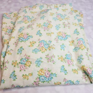 May include: A folded fabric sheet featuring an off-white background with a repeating floral pattern. The design consists of clusters of small roses in shades of light blue, soft yellow, and pale pink, accented with tiny green leaves and stems.