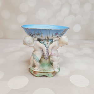 May include: A vintage porcelain bowl with cherub figures holding grapes. The bowl is light blue with gold accents and floral designs.