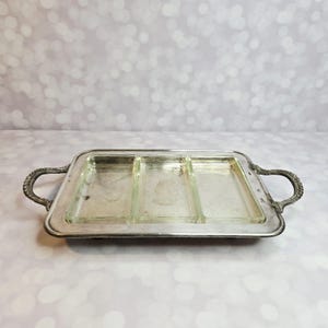 Puede incluir: Una bandeja rectangular plateada con tres compartimentos de vidrio. La bandeja tiene asas ornamentadas en cada extremo. Los compartimentos de vidrio son transparentes. La bandeja está diseñada para servir aperitivos o snacks.