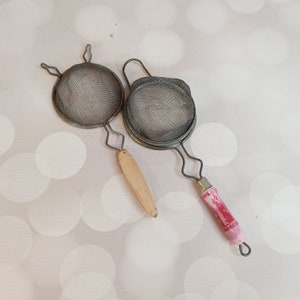 May include: Two metal mesh strainers with wooden and pink plastic handles. The strainers are round and have a fine mesh.