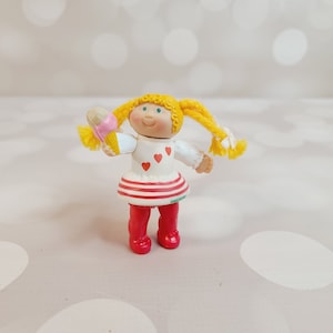 May include: A small plastic doll with blonde hair wearing a white dress with red hearts and red and white stripes, red shoes, and holding a pink and yellow ice cream cone.