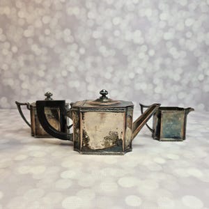 May include: A three-piece silver-plated tea set featuring a square-shaped teapot with a black handle, a matching square sugar bowl, and a square creamer. Each piece displays a tarnished metallic finish and intricate engraved border detailing.