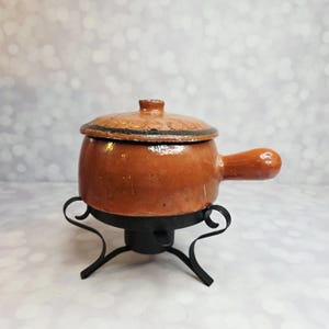 Puede incluir: Una pequeña olla de cerámica marrón con tapa y asa, colocada sobre un soporte de metal negro. La olla tiene un aspecto rústico, con un borde oscuro en la tapa. El soporte tiene un diseño curvo decorativo.