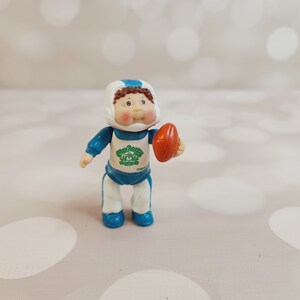 May include: A Cabbage Patch Kid doll wearing a blue and white football uniform with a white helmet and holding a brown football.