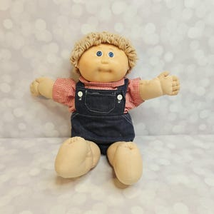 Puede incluir: Una muñeca de tela con piel bronceada clara, ojos azules y cabello de lana en tono marrón claro. La muñeca viste una camisa de cuadros rojos y blancos bajo un peto de mezclilla azul oscuro con dos botones blancos en los tirantes delanteros.