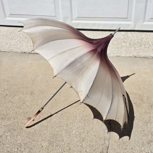 May include: An open, vintage-style umbrella with a unique, scalloped edge design. The umbrella features a gradient of colors, transitioning from a light cream to a deep burgundy at the center. The handle is a light brown color.
