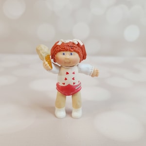May include: A red-haired Cabbage Patch Kid doll wearing a white shirt with red hearts and a red skirt. The doll is holding a small, white, heart-shaped object in her right hand.