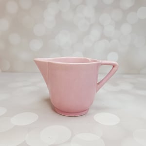 Puede incluir: Un jarro de crema de cerámica rosa con asa. El jarro está sobre una superficie blanca con un fondo borroso.