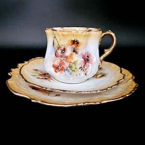 Peut inclure: Un service à thé vintage composé d'une tasse, d'une soucoupe et d'une assiette. L'ensemble est de couleur crème avec des motifs floraux peints à la main en rouge et orange. Les bords sont dorés, ajoutant une touche d'élégance.