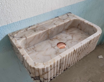 May include: A white marble sink with a copper drain stopper. The sink has a fluted edge and is mounted on a wall.