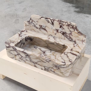 May include: A marble sink with a rectangular basin and a decorative backsplash. The sink features a cream-colored base with dark brown veining throughout. It is set on a wooden crate.