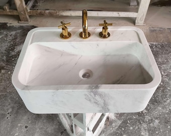 May include: A white marble bathroom sink with three gold faucets. The sink is rectangular and has a slightly curved edge. The sink is sitting on a wooden stand.
