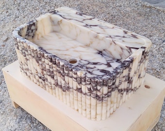 Può includere: Un lavabo in marmo bianco con venature viola e dorate. Il lavabo è circondato da un bordo decorativo di strisce di marmo verticali.
