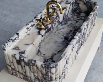 Könnte beinhalten: Ein weißes und violettes Marmor-Waschbecken mit geriffeltem Rand und zwei goldenen Wasserhähnen.