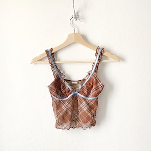 May include: A brown plaid camisole top with ruffled edges and light blue ribbon trim. The top is hanging on a wooden hanger against a white wall. The camisole has thin straps and a delicate, sheer fabric.