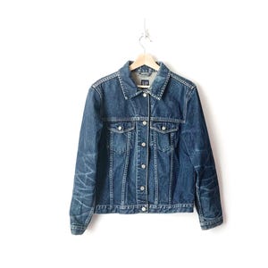 May include: A dark blue denim jacket with a button closure and two front pockets. The jacket has a faded look and is hanging on a wooden hanger.