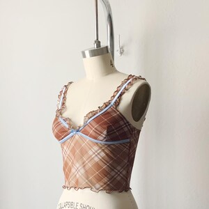 May include: A brown plaid crop top with a sheer overlay. The top features a ruffled trim along the neckline and straps, with a light blue accent. The garment is displayed on a mannequin.