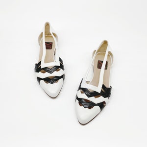 May include: A pair of white leather shoes with black leather cutouts. The shoes have a pointed toe and a low heel. The shoes are made by the brand 'Made in Italy'.