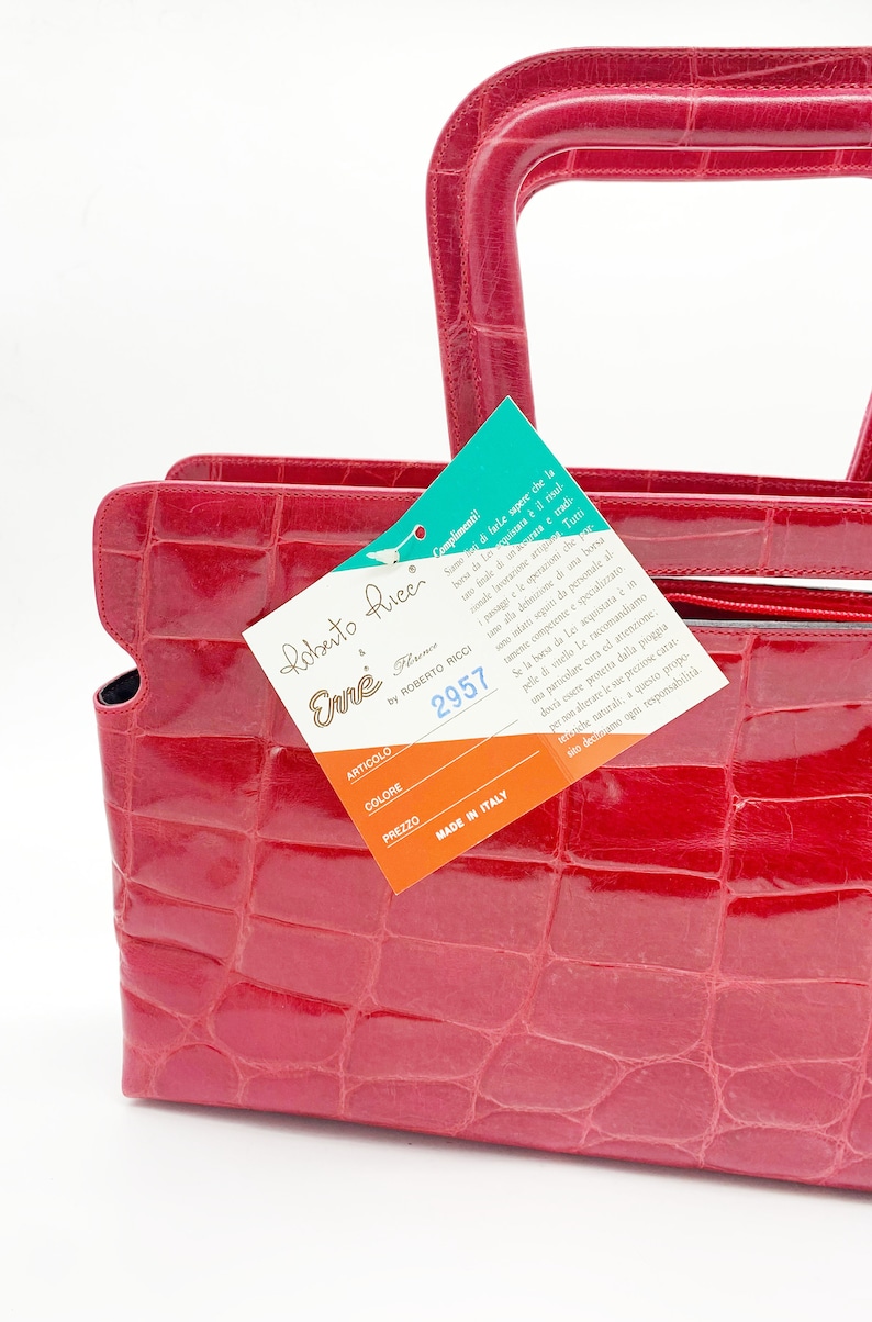 May include: A red crocodile embossed leather handbag with a top handle and a tag that reads "Roberto Ricci Enne Florence by Roberto Ricci 2957 Made in Italy".