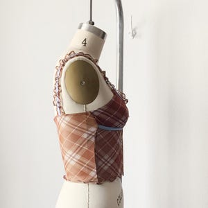 May include: A plaid corset top in shades of brown, tan, and burgundy, displayed on a mannequin. The top features a mix of plaid patterns, a ruffled trim, and a blue accent. The garment is designed with a fitted silhouette.