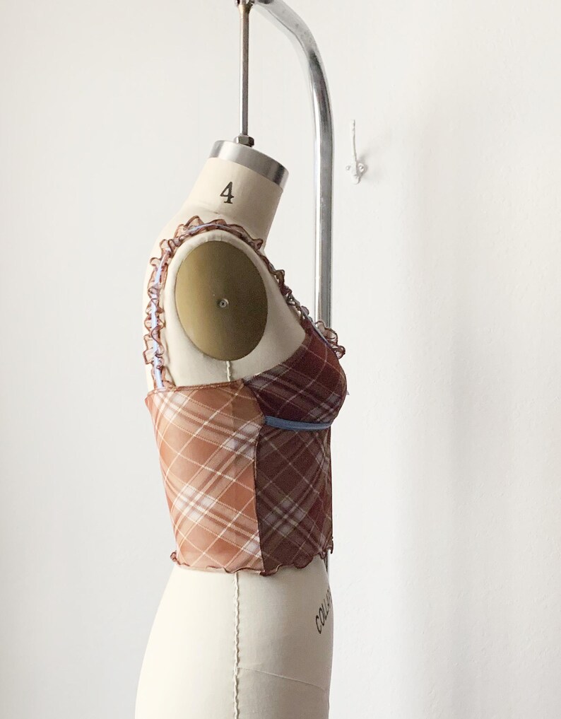 May include: A plaid corset top in shades of brown, tan, and burgundy, displayed on a mannequin. The top features a mix of plaid patterns, a ruffled trim, and a blue accent. The garment is designed with a fitted silhouette.