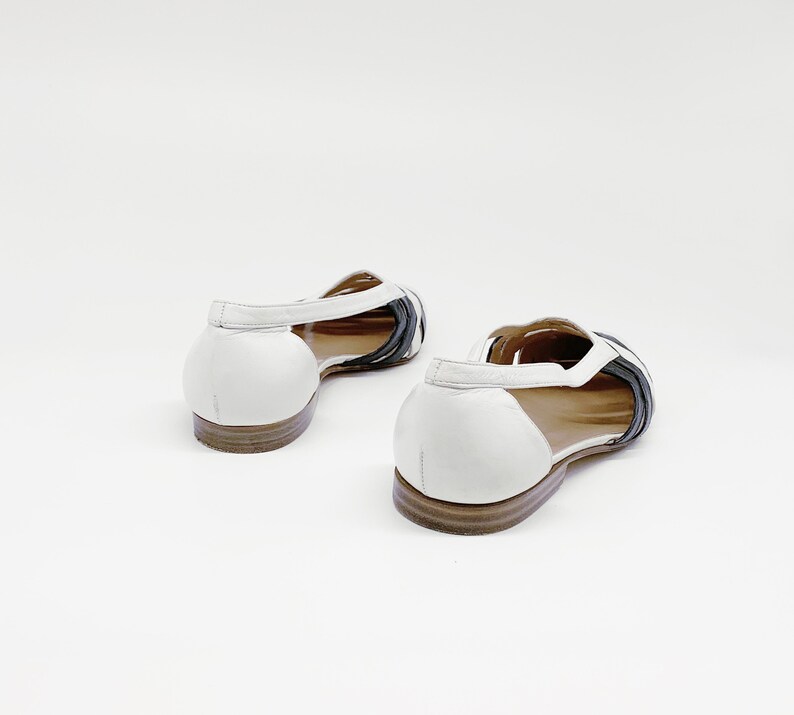 May include: A pair of white leather sandals with black leather straps. The sandals have a low heel and an open toe.