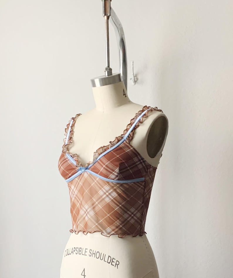 May include: A brown plaid crop top with a sheer overlay. The top features a ruffled trim along the neckline and straps, with a light blue accent. The garment is displayed on a mannequin.