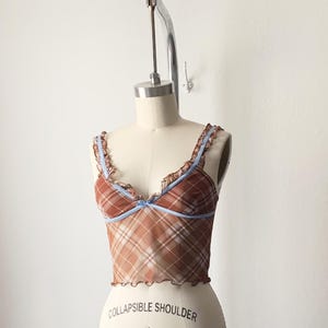 May include: A brown plaid camisole top with light blue trim and straps. The top has a ruffled edge and a small bow detail at the center. The garment is displayed on a mannequin.