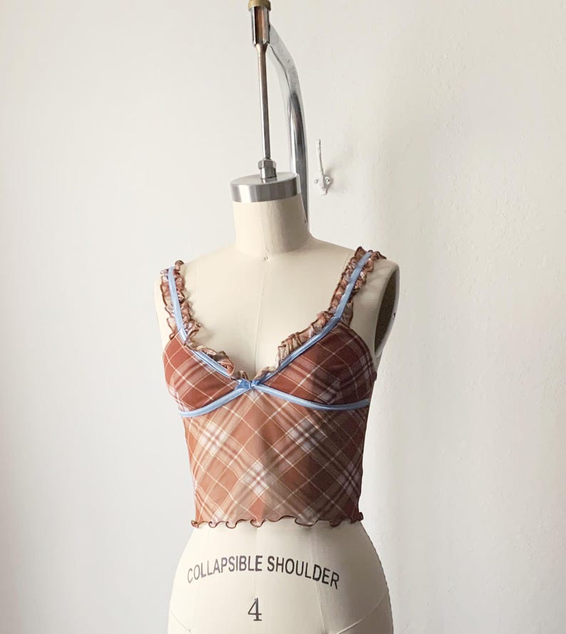 May include: A brown plaid camisole top with light blue trim and straps. The top has a ruffled edge and a small bow detail at the center. The garment is displayed on a mannequin.