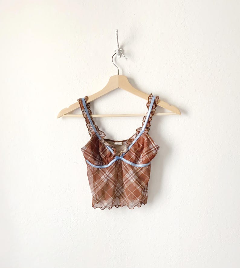 May include: A brown plaid camisole top with ruffled edges and light blue ribbon trim. The top is hanging on a wooden hanger against a white wall. The camisole has thin straps and a delicate, sheer fabric.