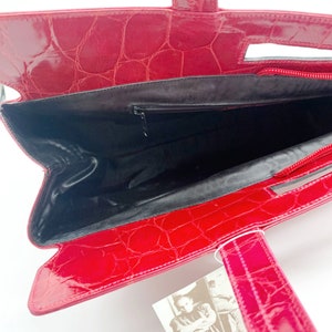 May include: Red crocodile embossed leather handbag with a black interior and a zipper closure. The bag has a top handle and two shorter handles. The bag has a tag with the text "Roberto Ricci" and contact information.
