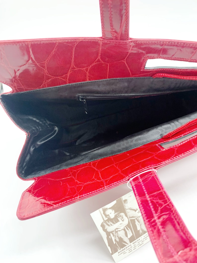 May include: Red crocodile embossed leather handbag with a black interior and a zipper closure. The bag has a top handle and two shorter handles. The bag has a tag with the text "Roberto Ricci" and contact information.