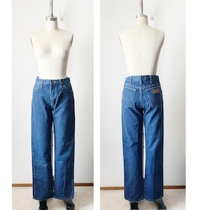 May include: A pair of blue denim jeans with a straight leg fit. The jeans have a brown leather patch on the back with the brand name "Wrangler" written on it.