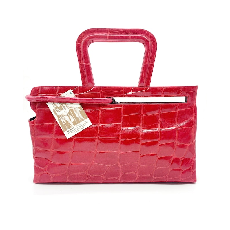 May include: A bright red crocodile embossed leather handbag with a unique top handle. The bag has a small tag attached with the text "Roberto Ricci, Made in Italy, Genuine Leather, Art. 1001".