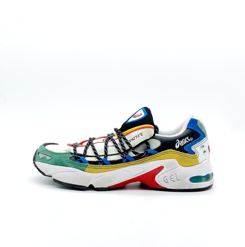 May include: White, green, blue, yellow, and red sneakers with black laces and a black and red logo that says "asics" on the side. The sneakers have a white sole with a red "gel" logo on the heel.