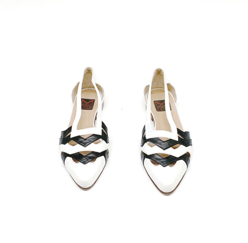 May include: A pair of white and black leather sandals with a cut-out design. The sandals have a pointed toe and a low heel.