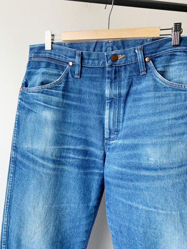 May include: A pair of blue denim jeans with a wooden hanger. The jeans have a classic five-pocket style and a straight leg.